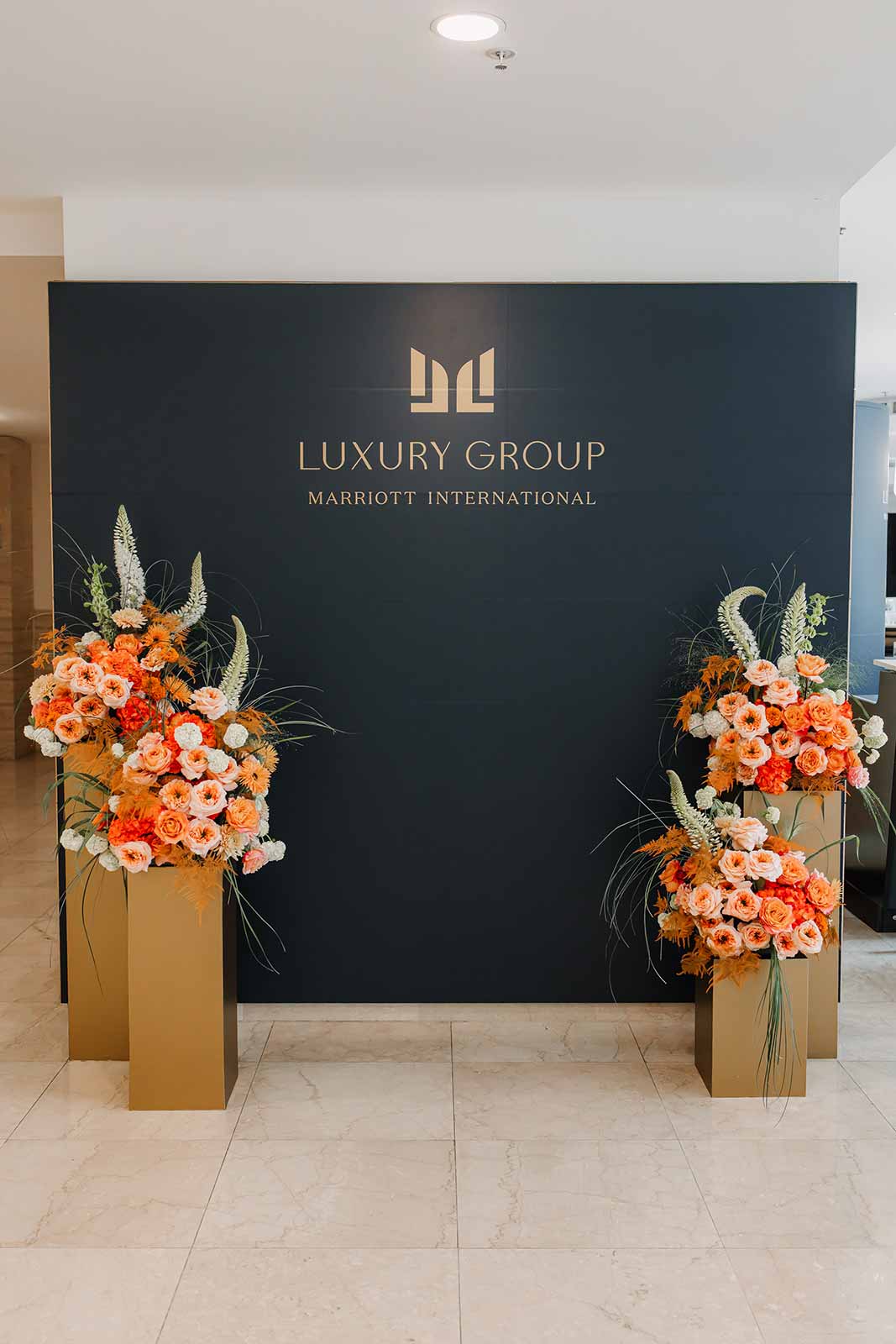 Navy wall wrap with a gold Marriott Luxury Group logo, framed by two orange floral arrangements